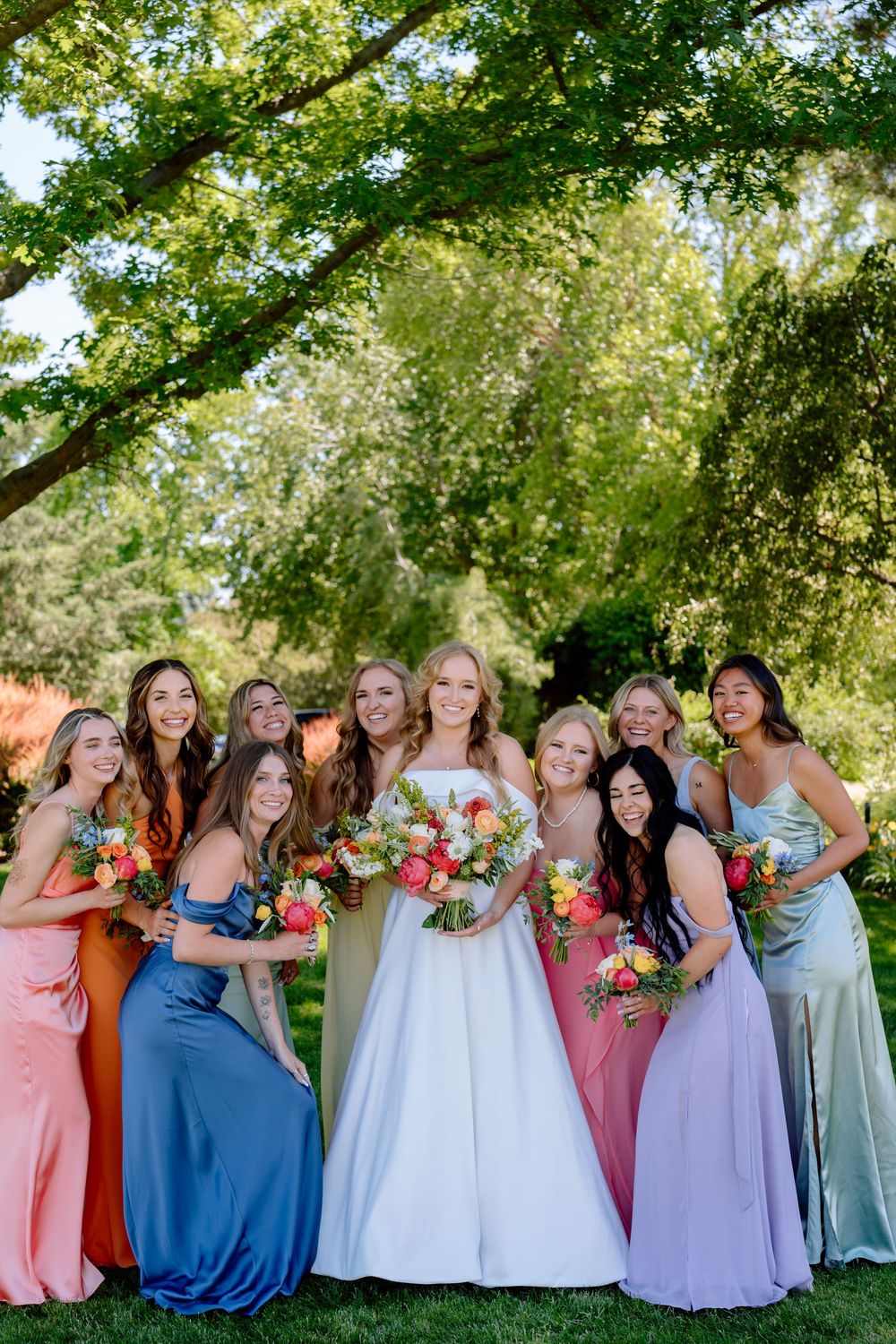 A group in colorful dresses poses together at an outdoor event under green trees.