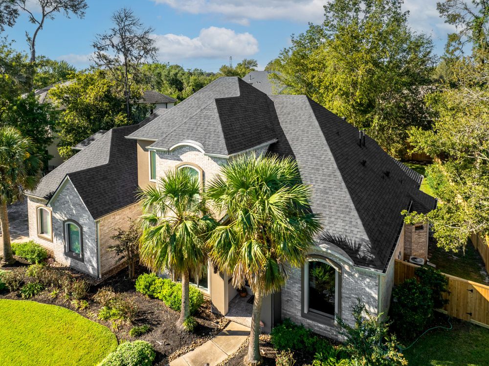 Luxurious two-story brick home with black roof and palm tree in well-landscaped front yard viewed from above.