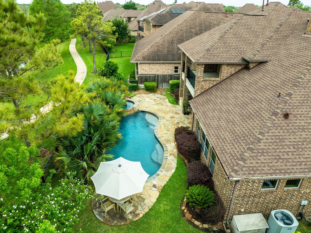 Aerial view of a luxury brick home with a freeform pool, stone patio, umbrella seating, and lush landscaping.