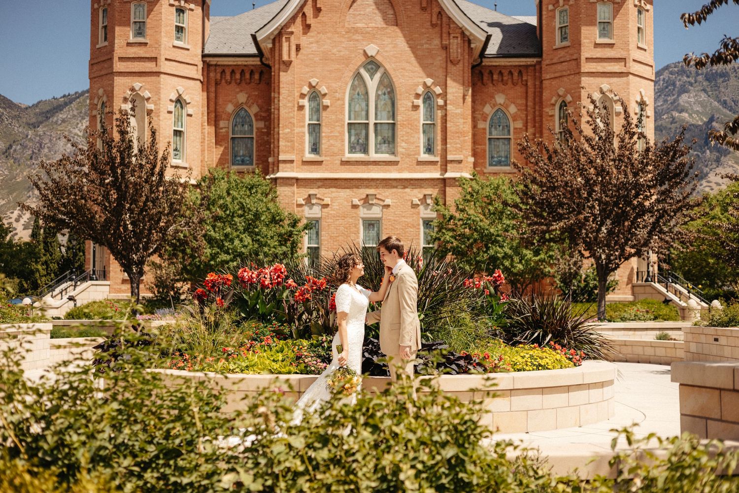 A couple poses in wedding attire in front of a historic red brick church with mountain backdrop.