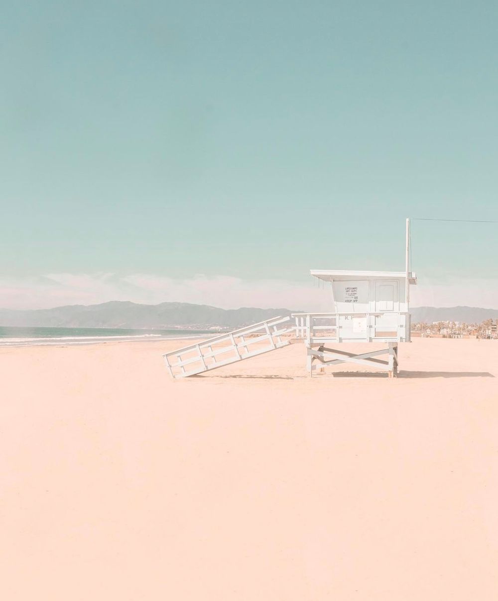 Minimalist beach scene with white lifeguard tower on pink sand against pale blue sky.