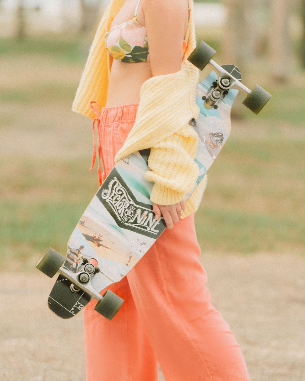 Person wearing coral pants holding a longboard skateboard with white and mint green design against a blurred outdoor background.