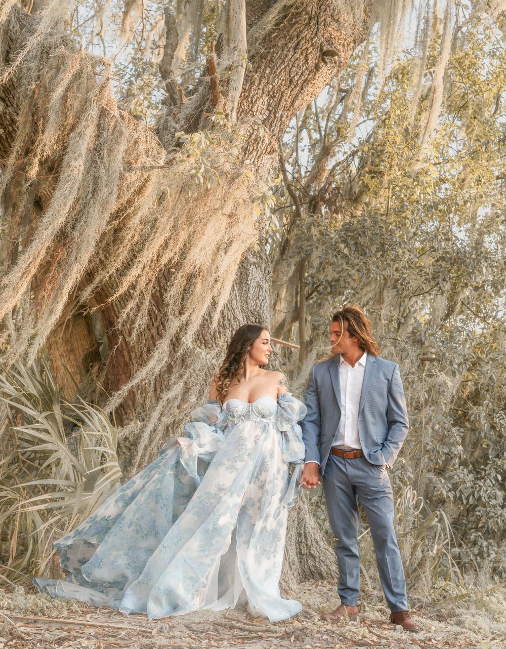 A romantic couple poses in ethereal attire against a dramatic rock wall backdrop in soft natural lighting.