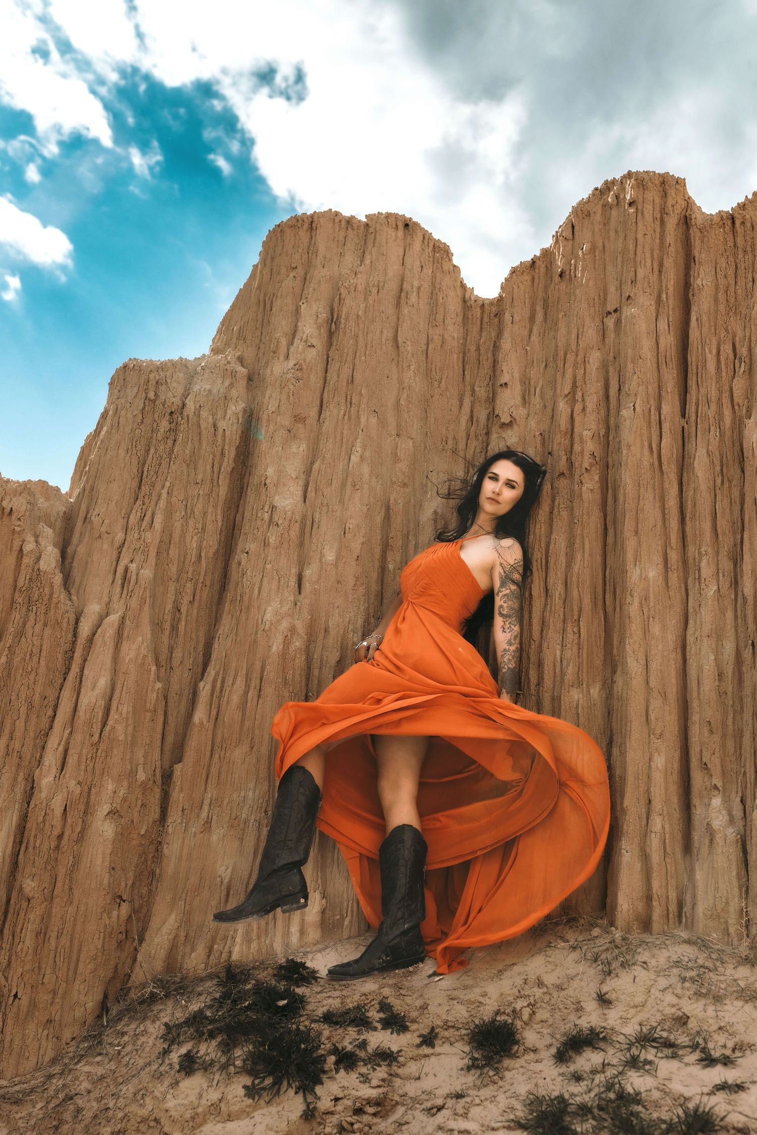 Woman in flowing orange dress and black boots poses against tall sandy rock formations under a blue sky.