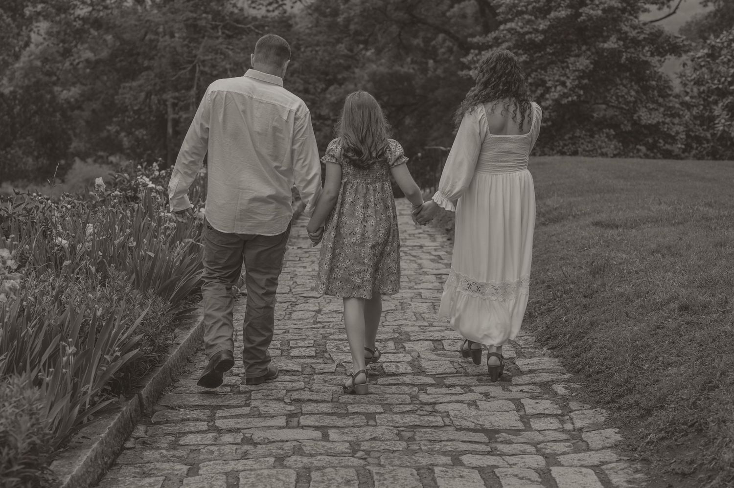 A black and white photo by samantha cambell imagery shows a family walking away on a cobblestone path through a garden landscape.