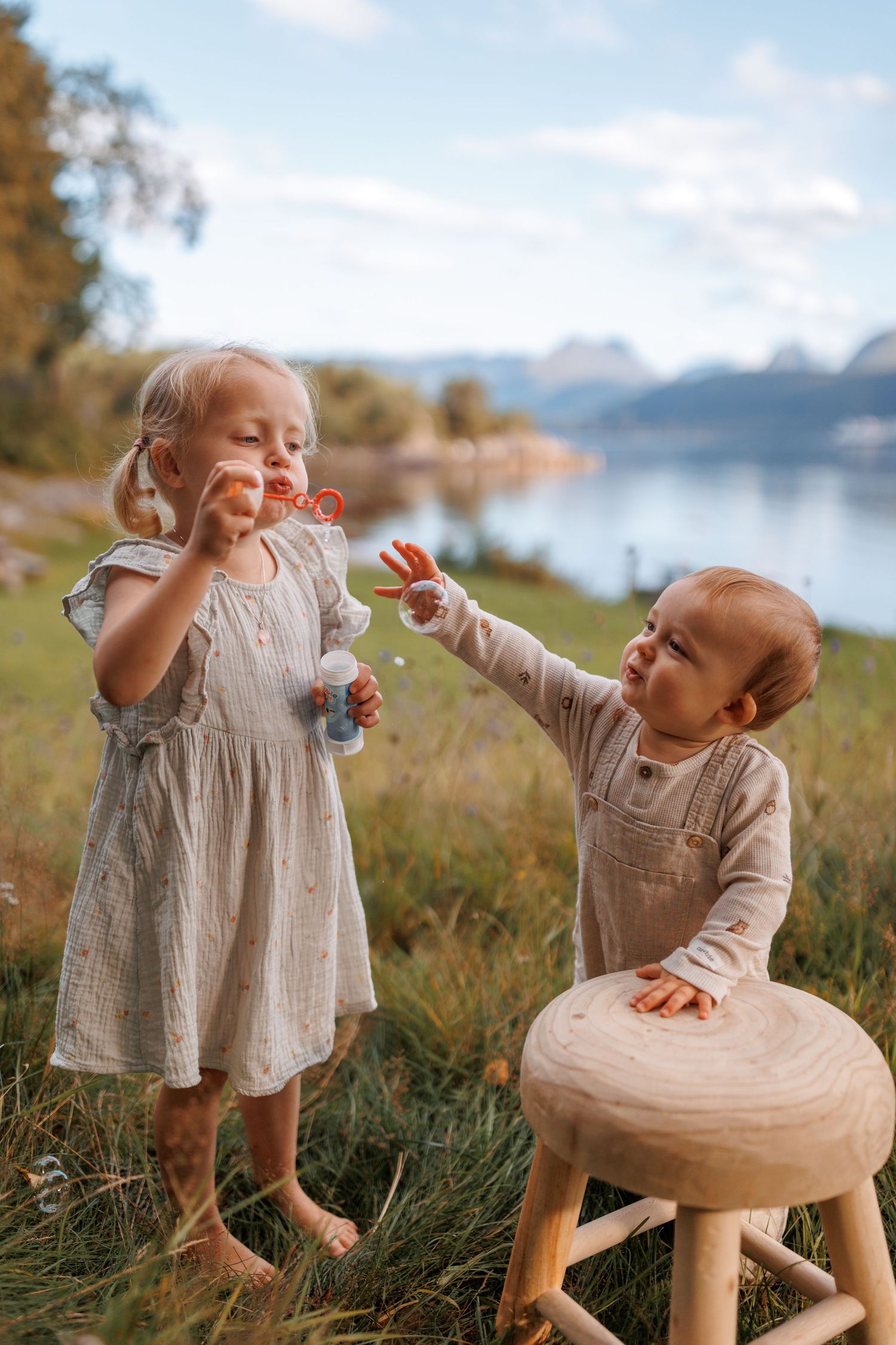 Two children playing with bubbles by a lake on a sunny day, with mountains in the background.