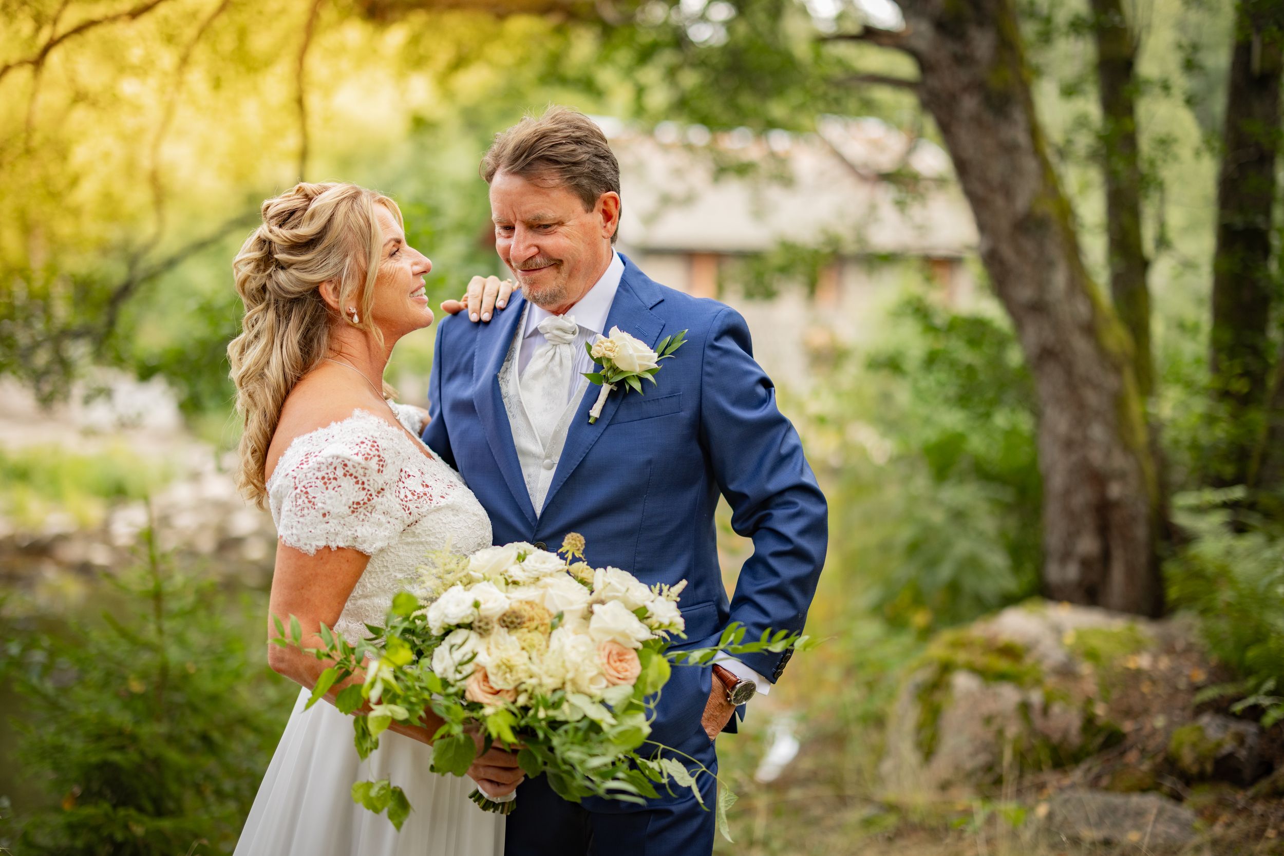 Bride and groom share a romantic moment outdoors, she holds a white bouquet, he wears a blue suit, surrounded by lush greenery.