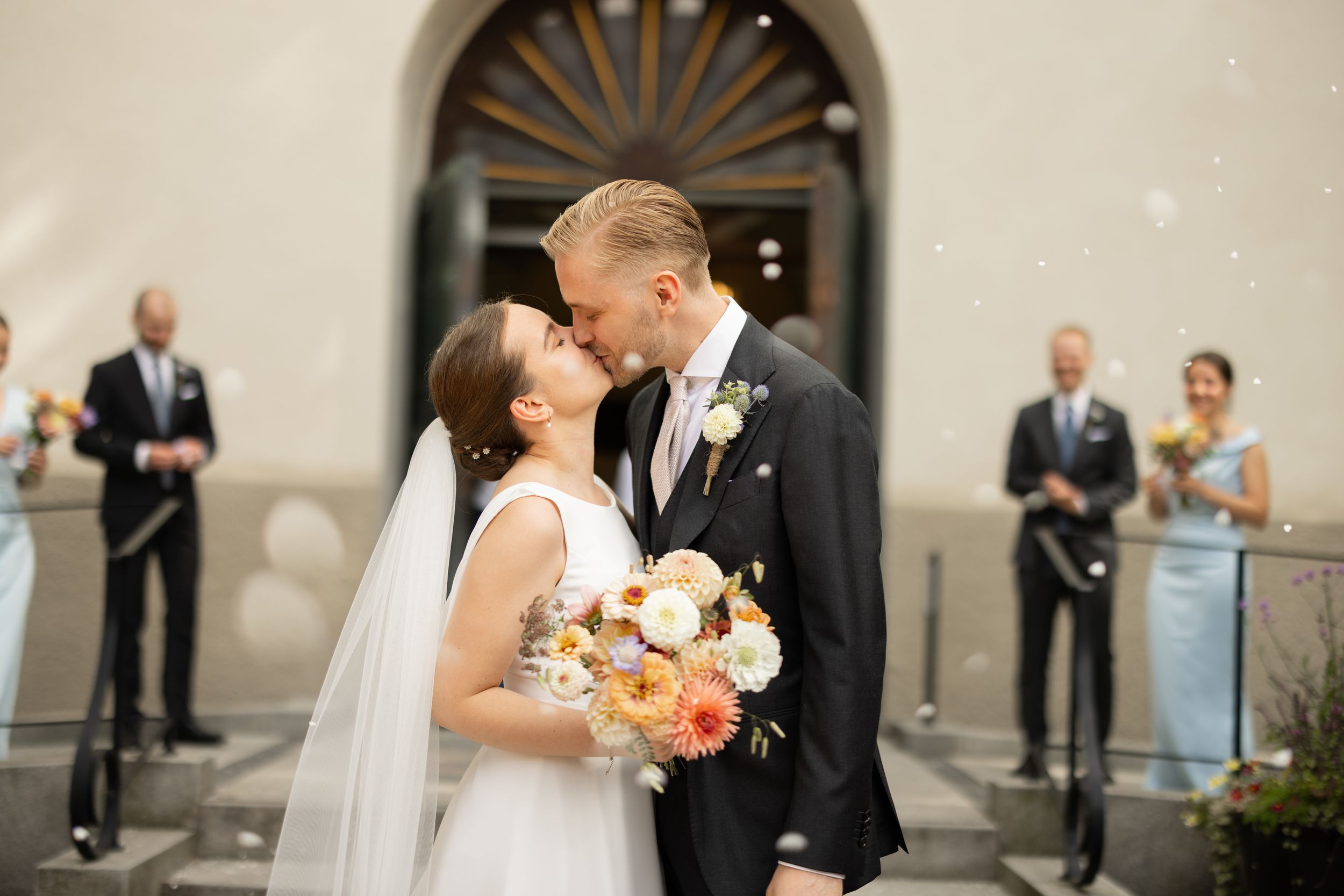 Bride and groom share first kiss outside church while guests celebrate with confetti and bouquet of orange flowers.