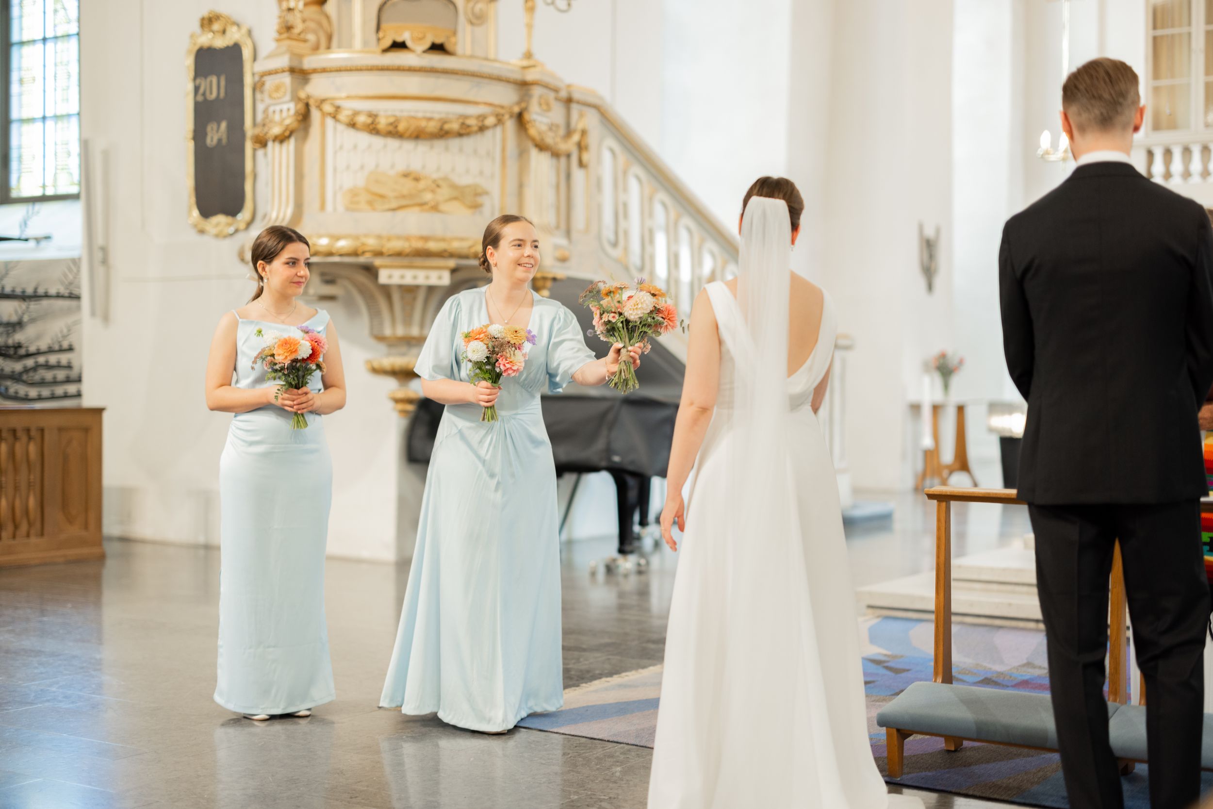 Bride in white gown stands with bridesmaids in light blue dresses holding bouquets inside an elegant church.