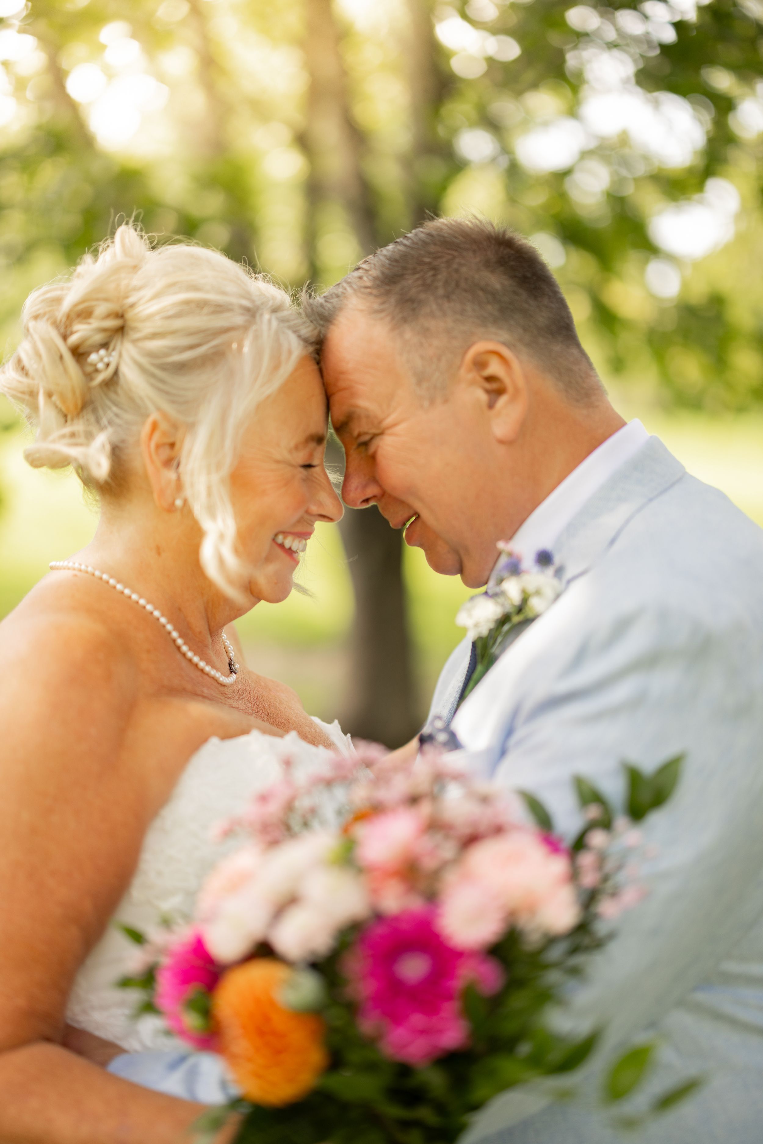 Smiling mature bride and groom touch foreheads tenderly outdoors, holding colorful wedding bouquet with pink and orange flowers.