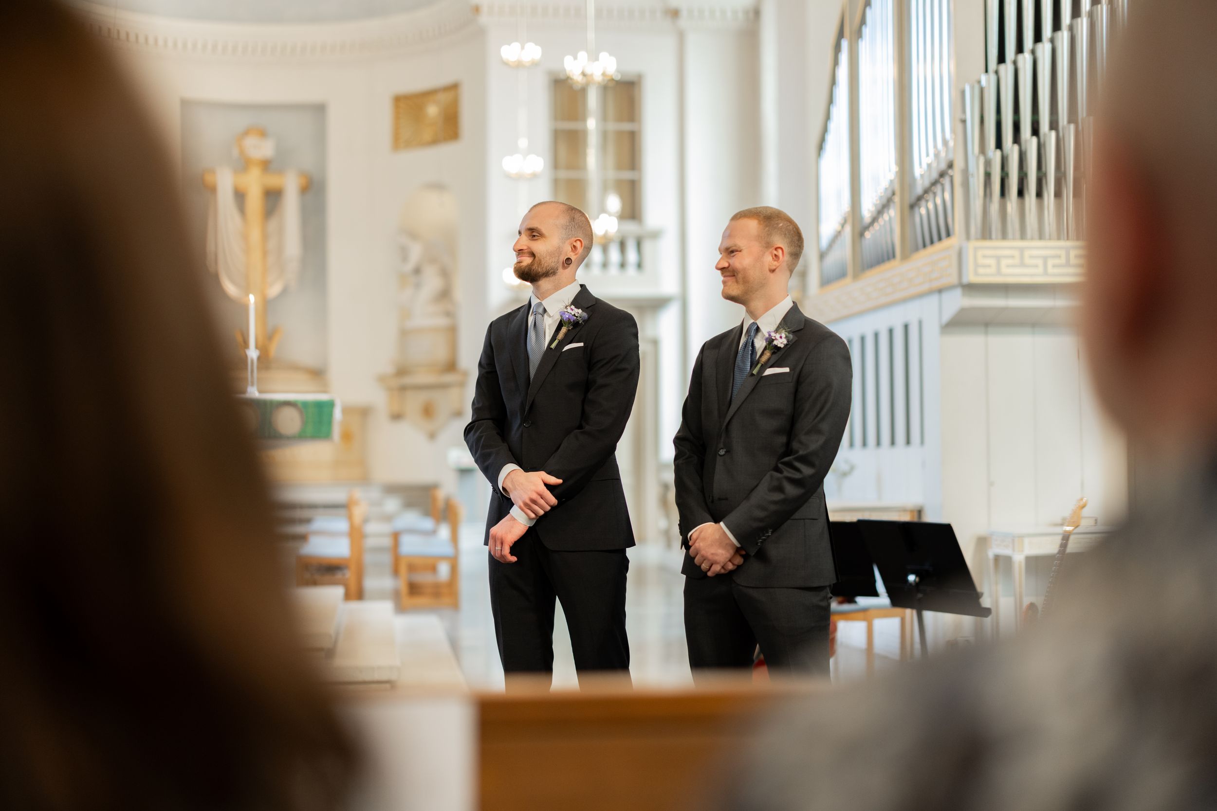 Two grooms in dark suits smiling at the altar inside an elegant white church during their wedding ceremony.