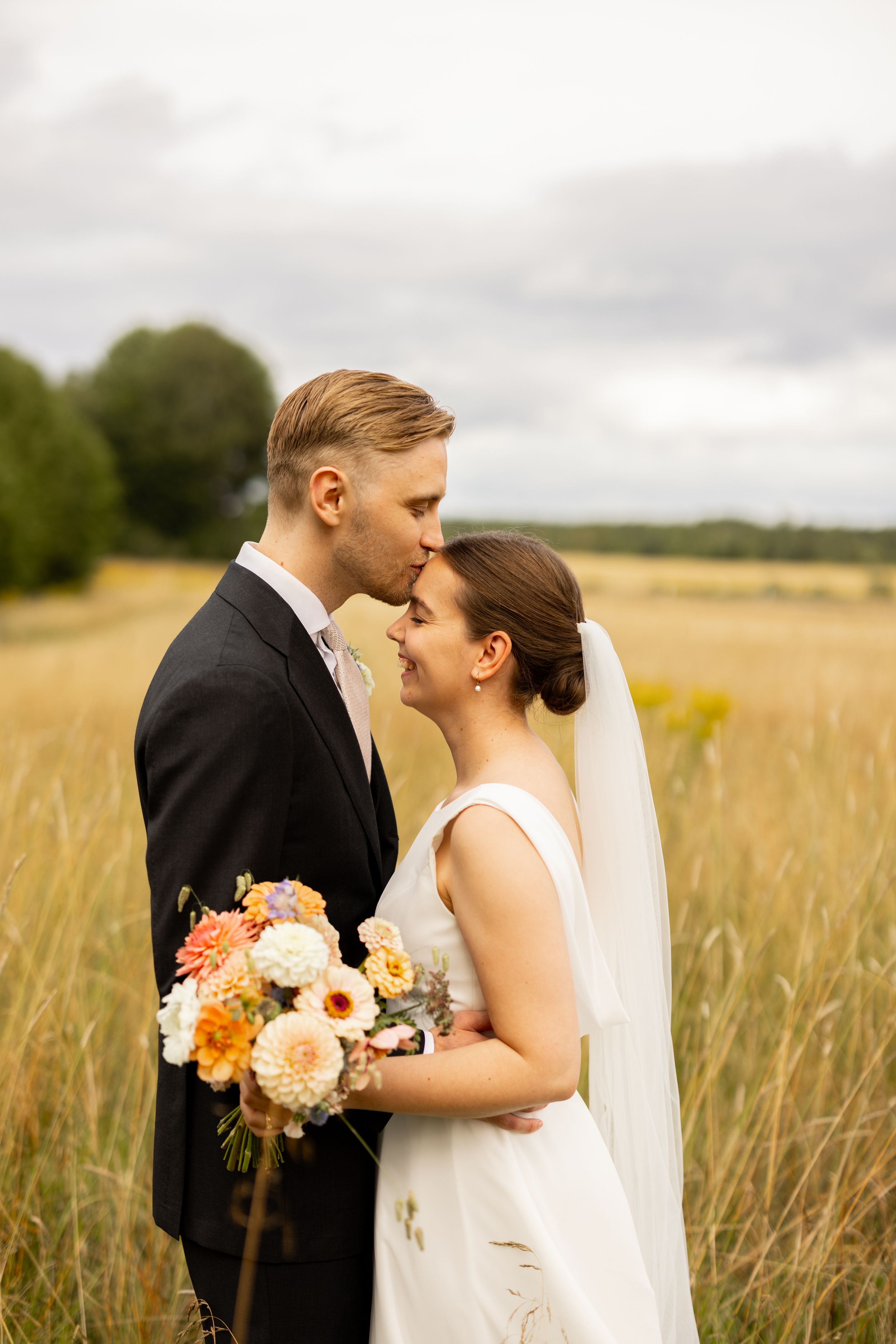 Bride and groom share a tender kiss in a golden wheat field, bride holding a colorful bouquet in white gown.