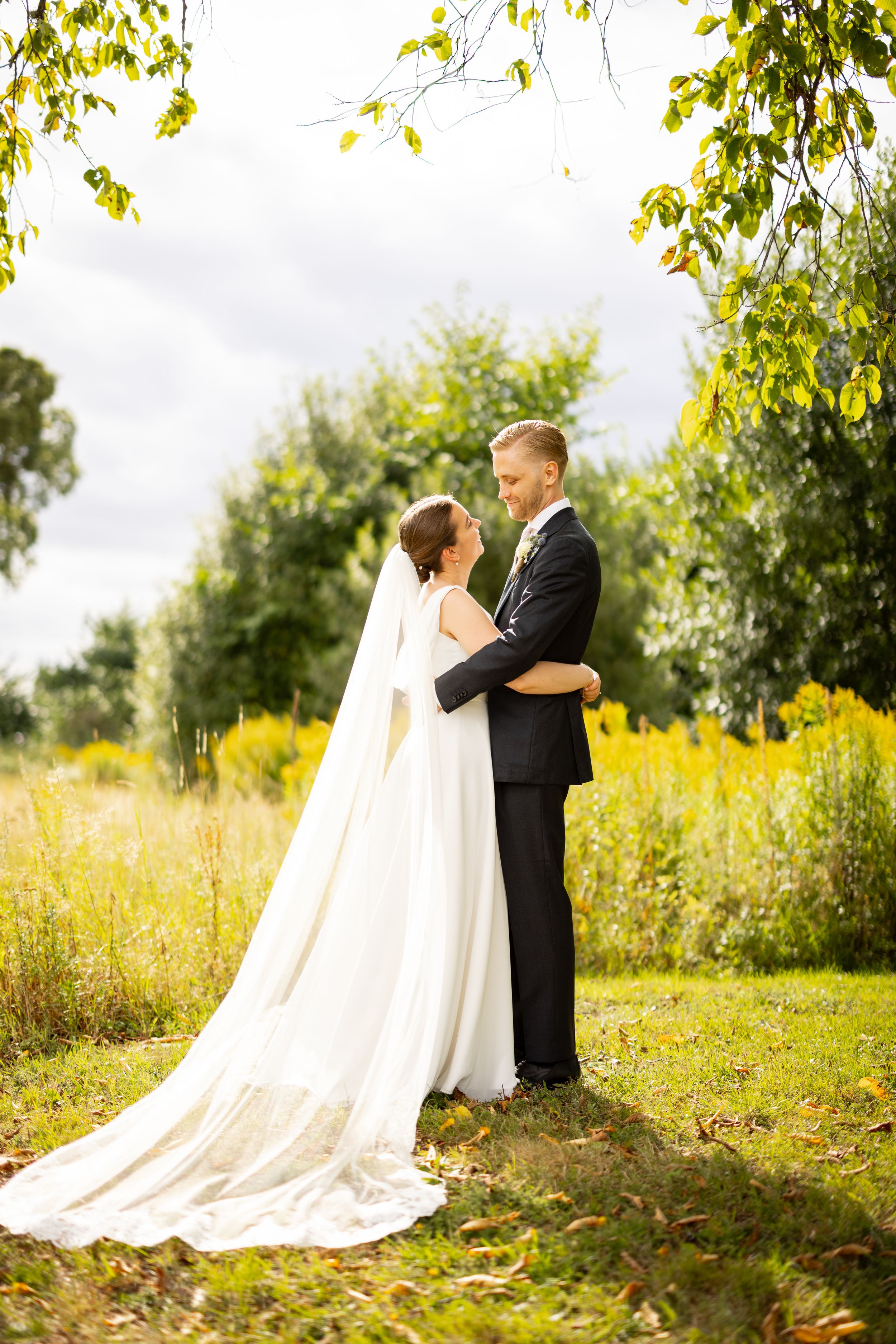 A bride and groom embrace outdoors in a lush green field, the bride wearing a flowing white gown with a long veil.