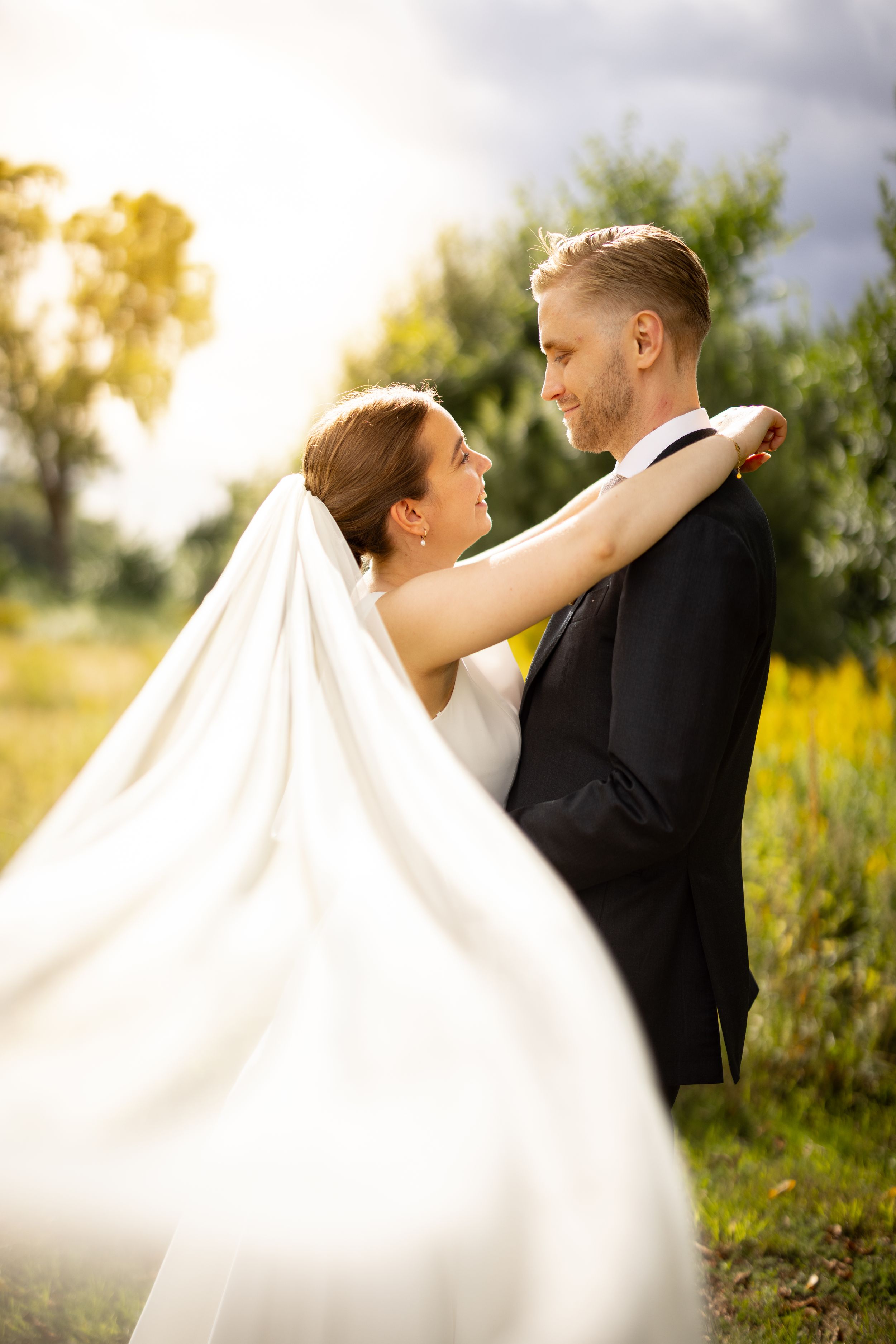 Bride and groom embrace outdoors in golden sunlight, her white veil flowing, surrounded by lush greenery.