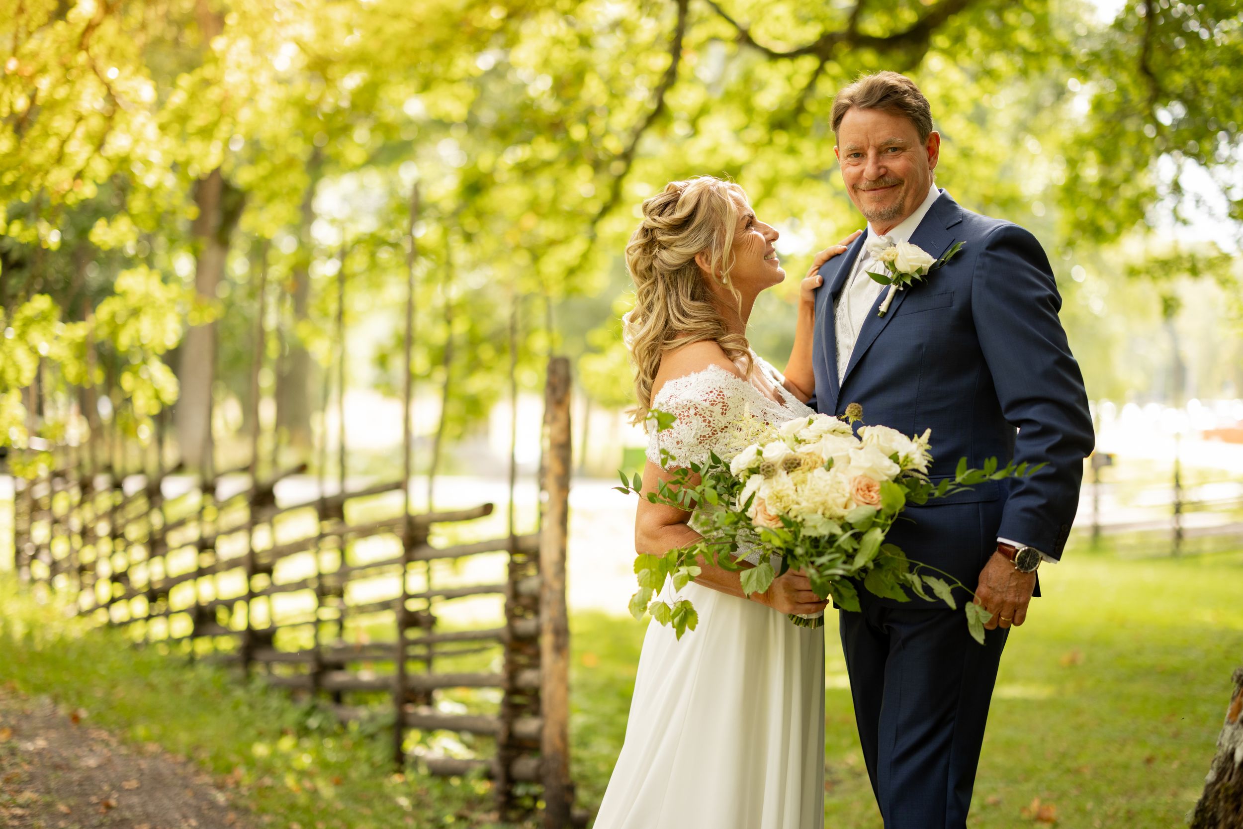 Bride and groom pose outdoors near a wooden fence surrounded by lush green trees on their wedding day.
