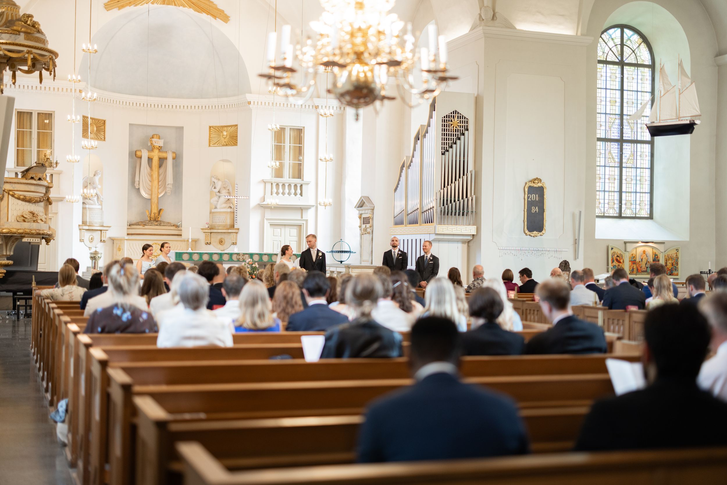 Guests seated in wooden pews during a wedding ceremony inside a bright, ornate white church with chandelier.