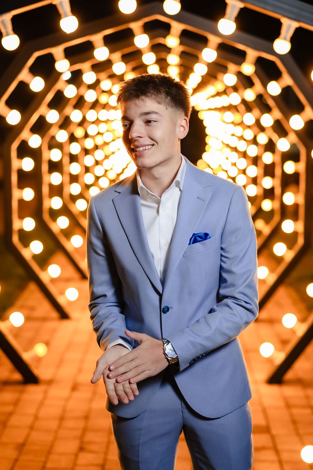 Stylish portrait against glowing string lights backdrop wearing light blue suit and white shirt.