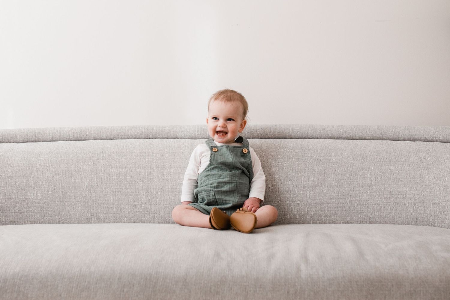 Adorable smiling baby in green overalls sits cross-legged on a light gray couch against a white wall.