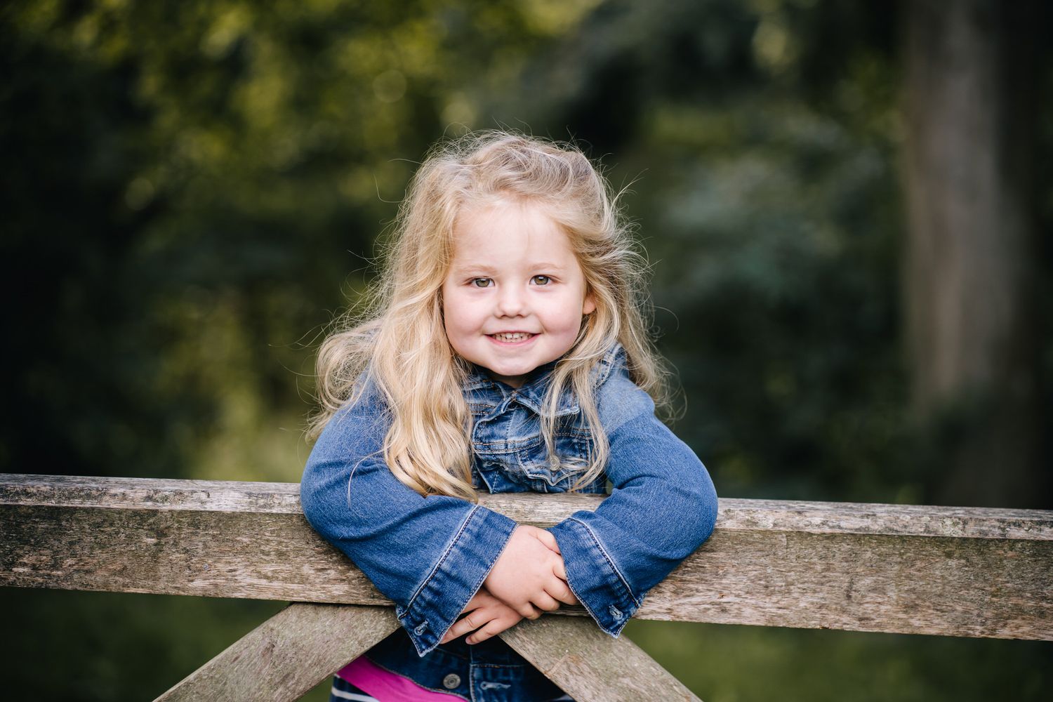 A smiling young girl in a fur-trimmed vest leans on a wooden fence surrounded by autumn foliage.