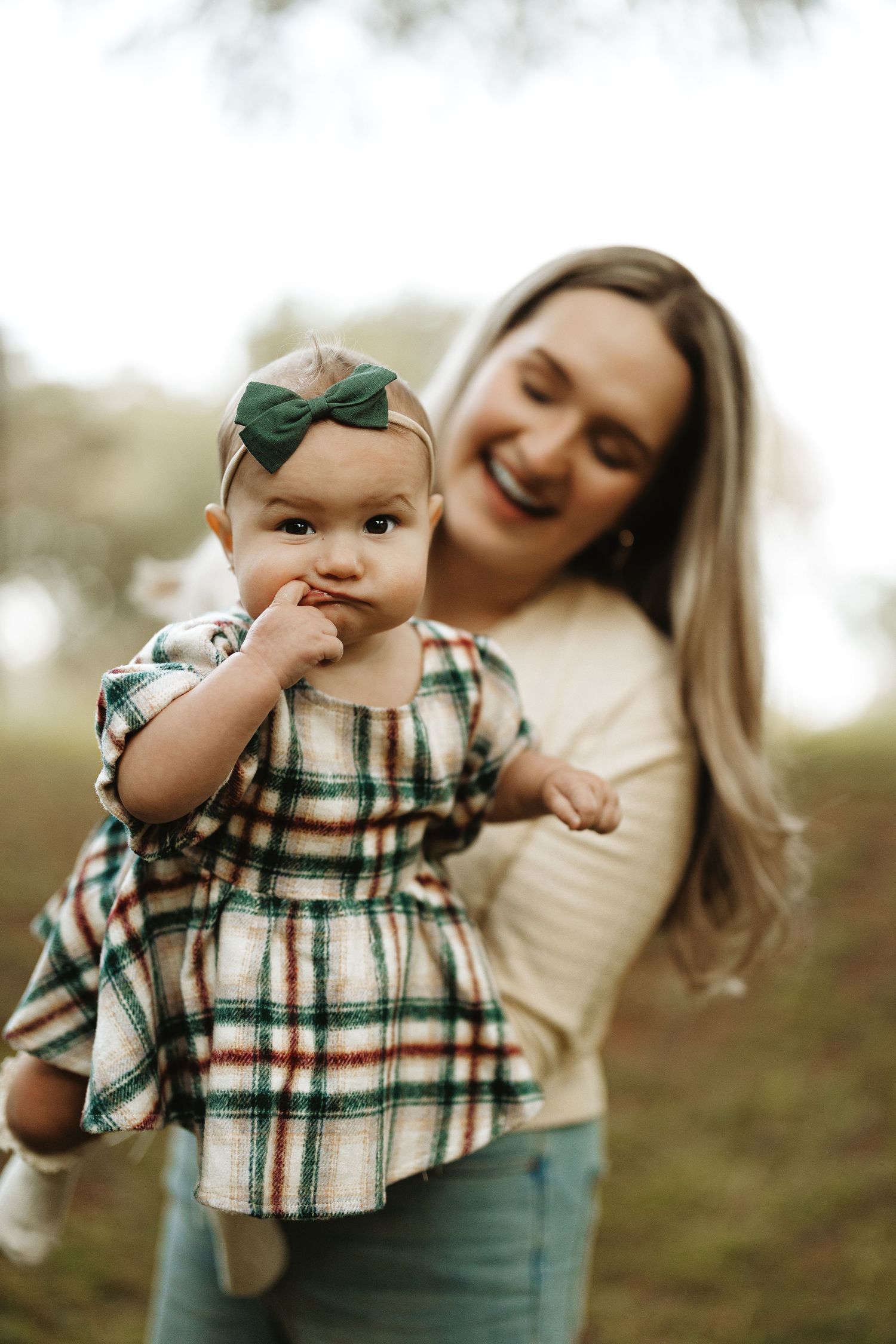 A baby in plaid dress smiles while being held by their parent in a natural outdoor setting.