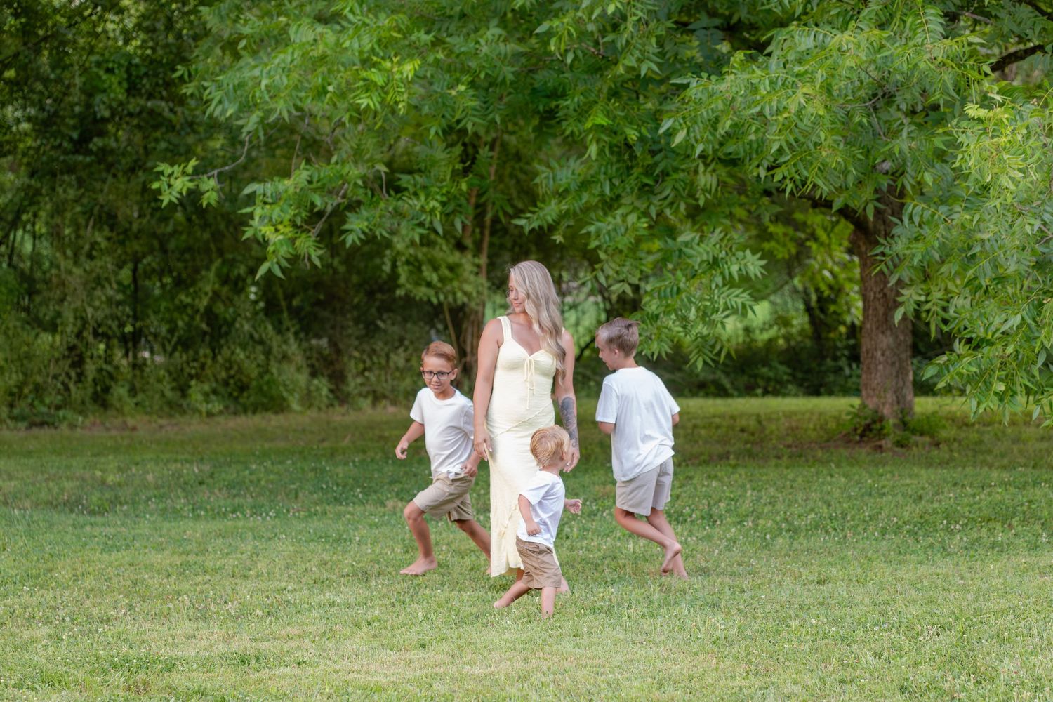family photographer Calhoun, GA A mother in a yellow dress walks barefoot on green grass with three young children in a sunny field.