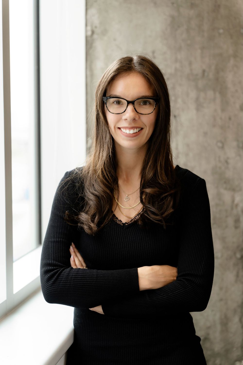 Person in black sweater and glasses poses confidently by window with industrial concrete background.
