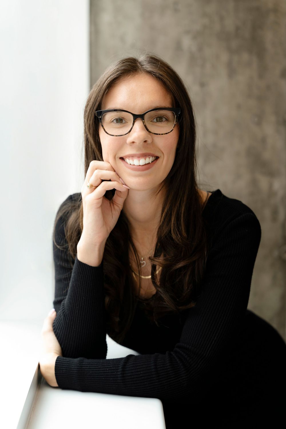 Person in black sweater and glasses smiles warmly while resting chin on hand.