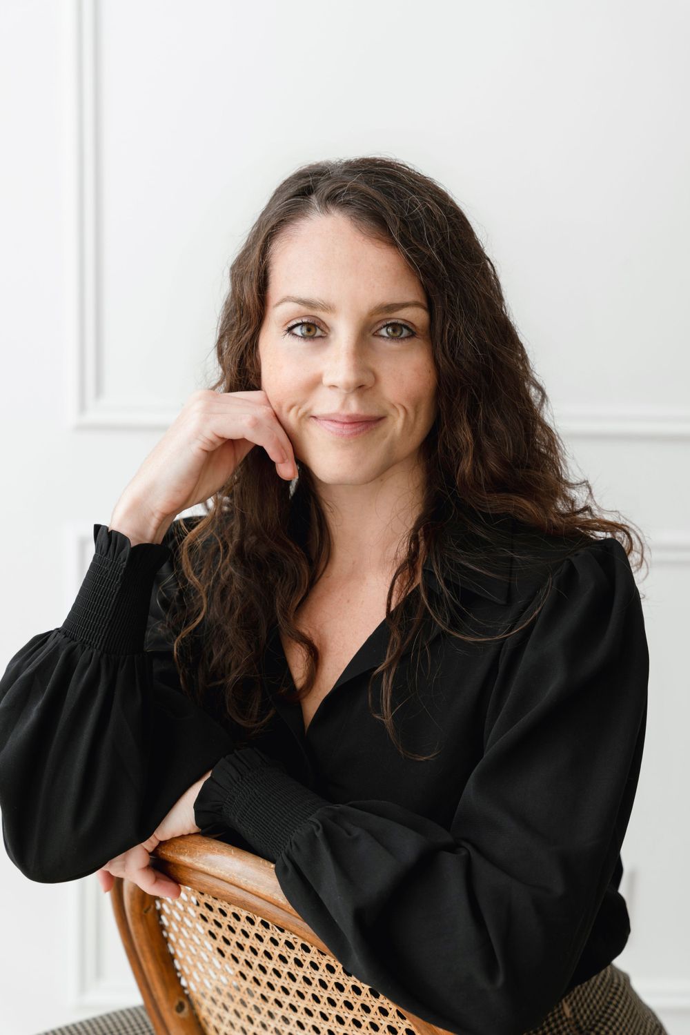 Professional portrait of woman in black blouse sitting on cane chair against white wall.