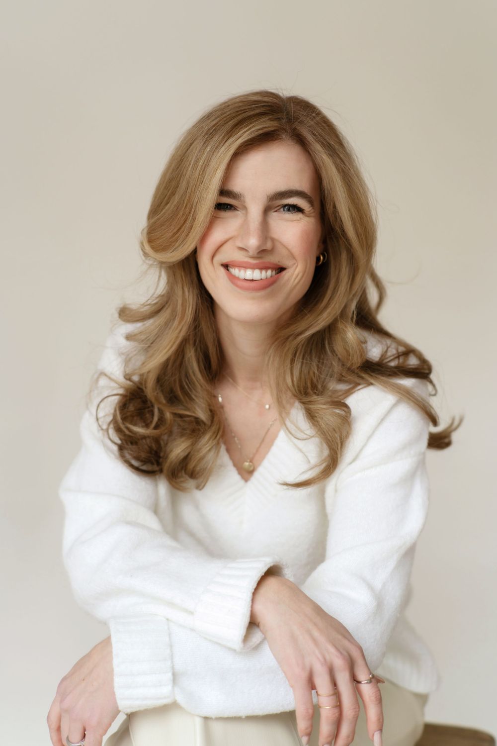 Smiling woman in white sweater with styled blonde hair against light backdrop.