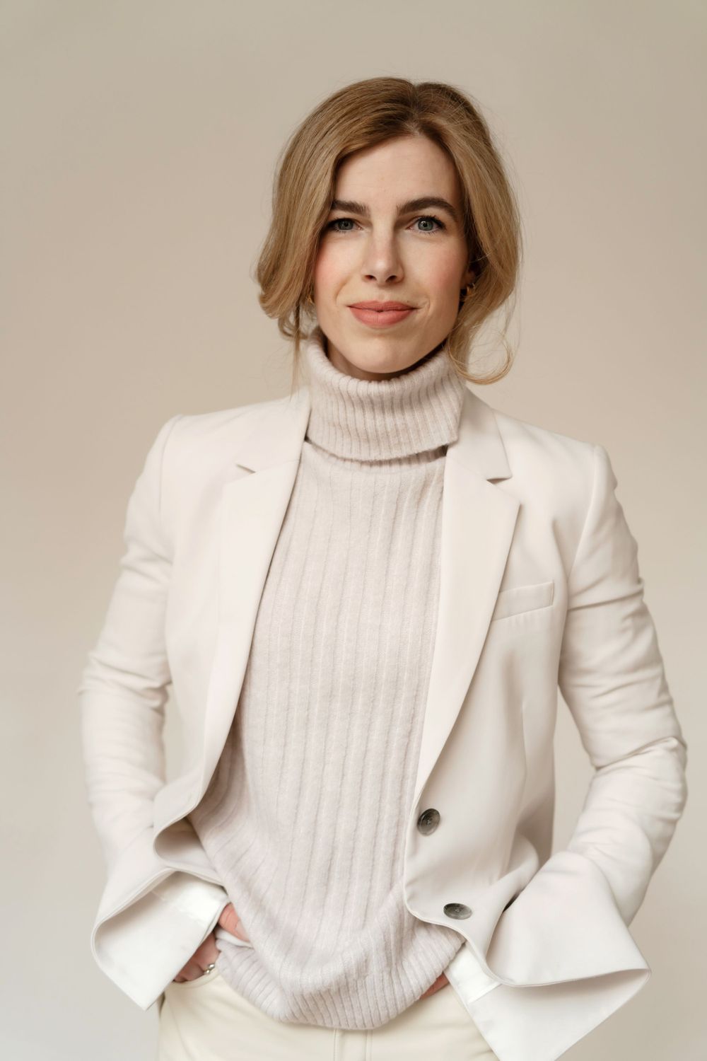 Woman in cream turtleneck and white blazer poses confidently against neutral background.
