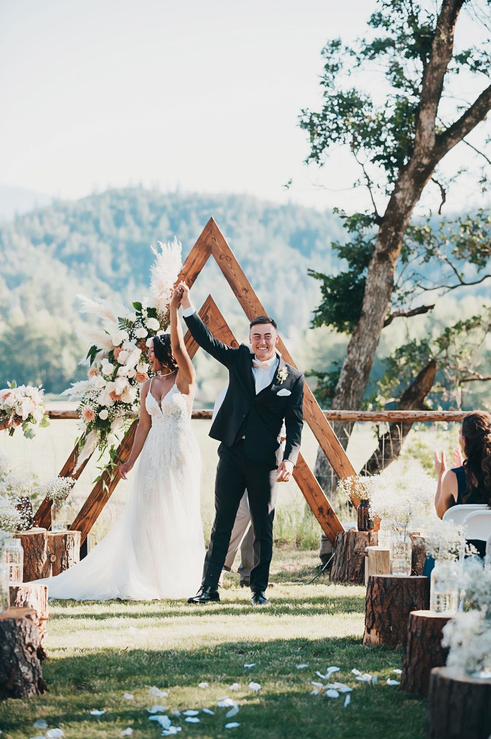 A wedding ceremony setup in a mountain meadow with wooden triangle arches decorated with flowers and greenery.