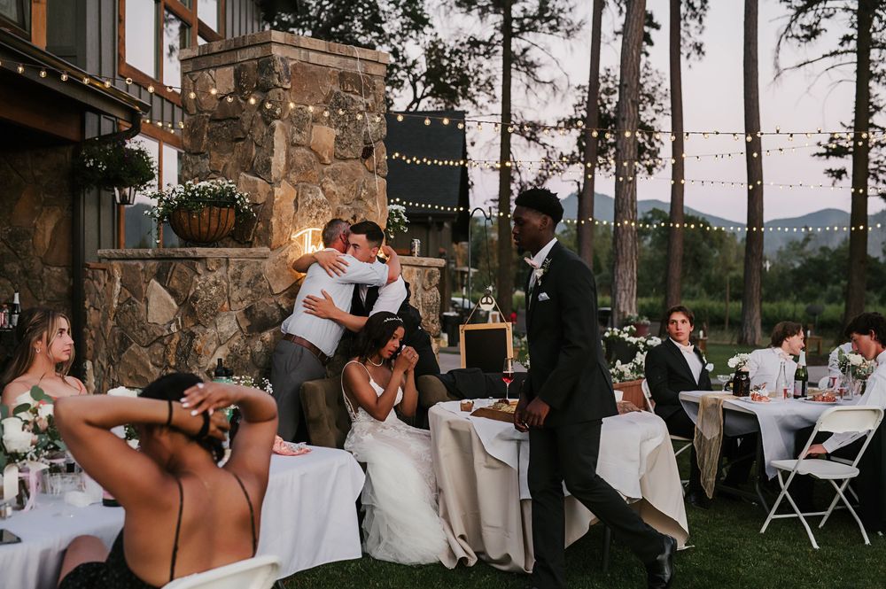 Wedding guests enjoy an outdoor reception dinner at sunset with tall pine trees and white tablecloths in view.
