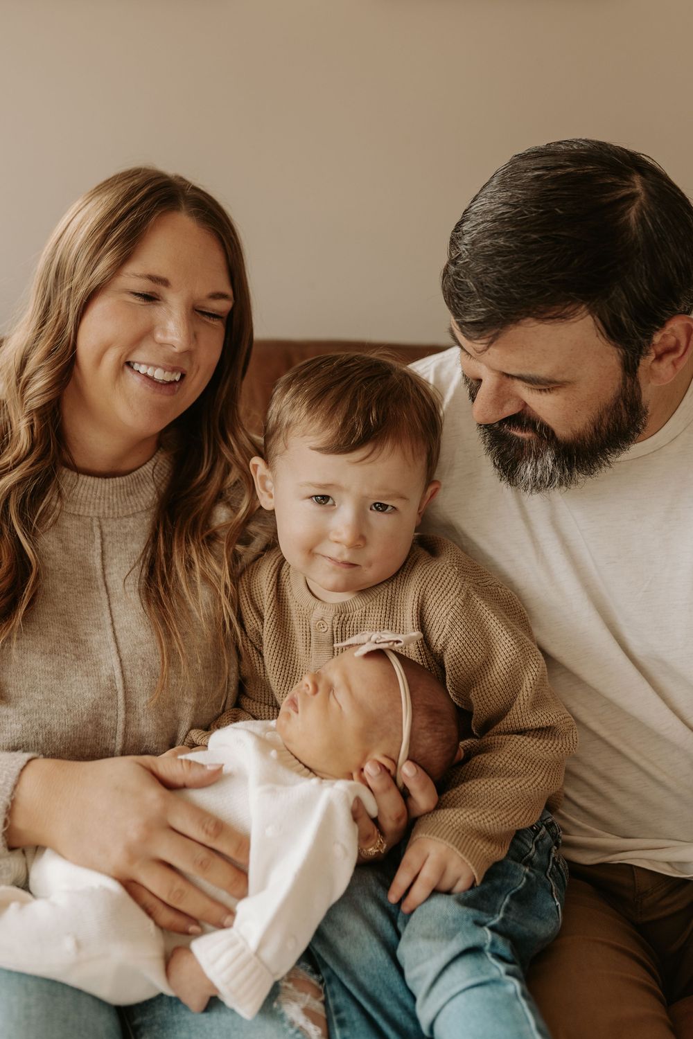 Happy family portrait with parents and toddler admiring their newborn baby while sitting together on a couch.