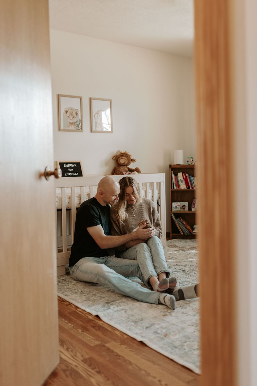 Couple sitting on nursery floor holding newborn baby, with white crib and animal decor in a cozy room.