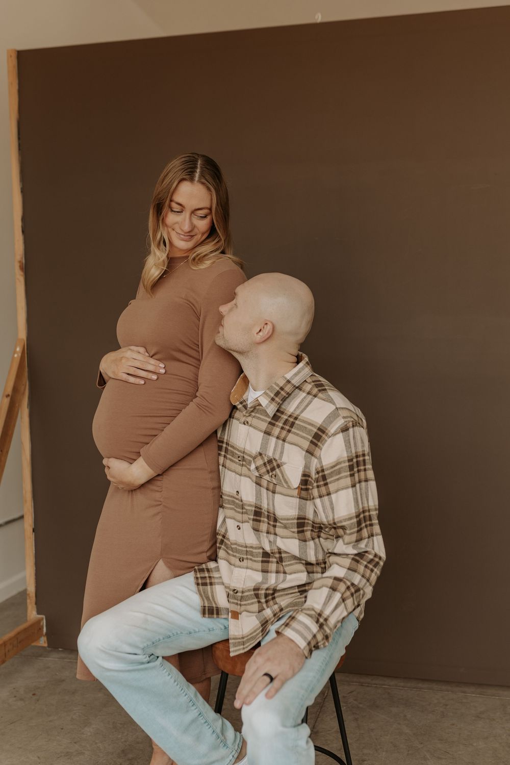Pregnant woman in brown dress with partner kissing her baby bump in front of a dark brown backdrop.