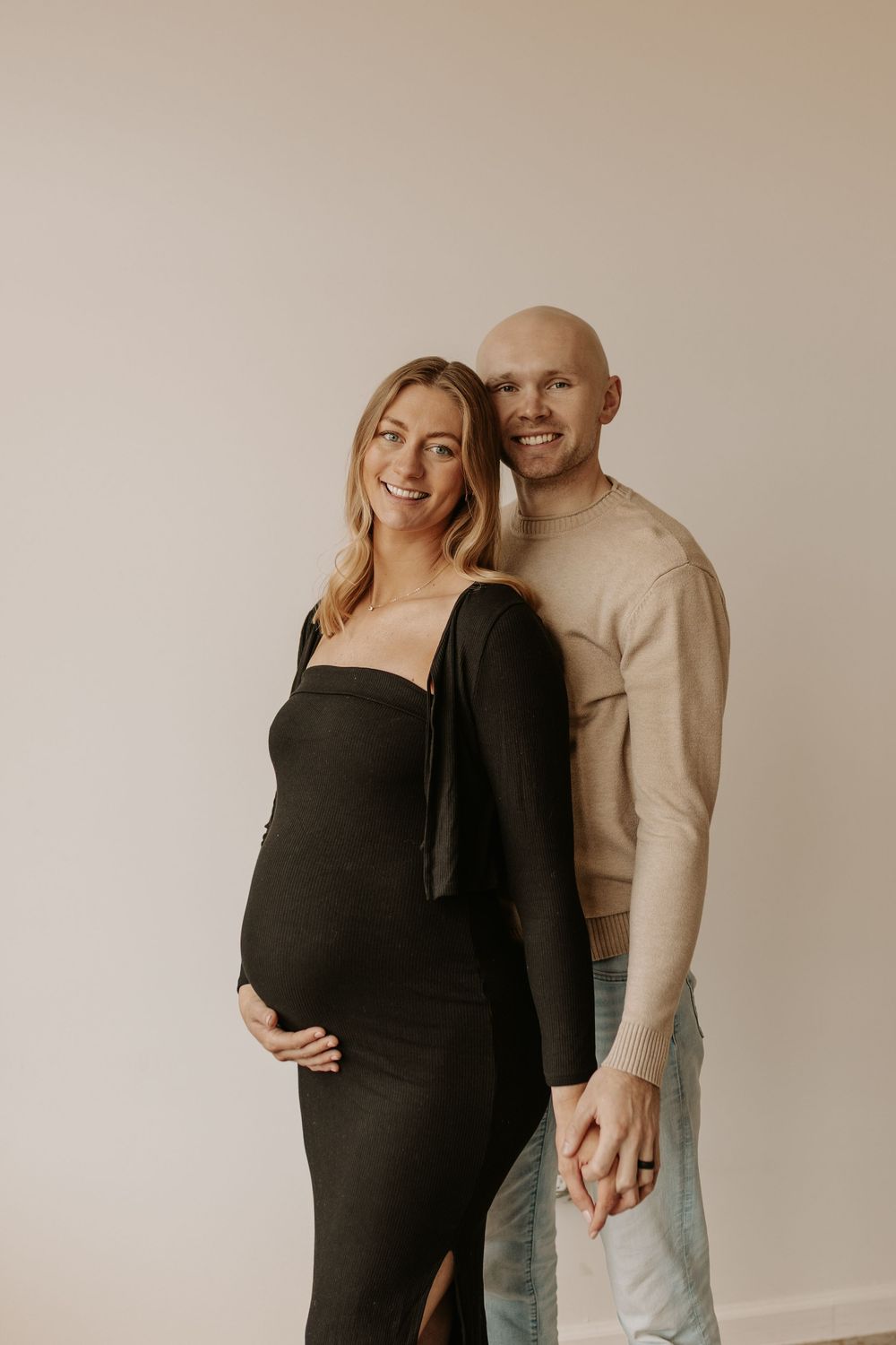 Smiling pregnant woman in black maternity dress with bald partner in beige sweater posing together against white wall.
