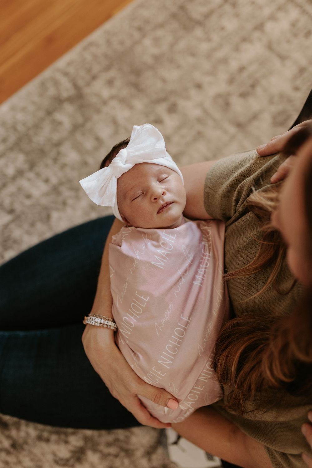 Newborn baby wearing pink outfit being held and kissed by parents on a beige carpet.