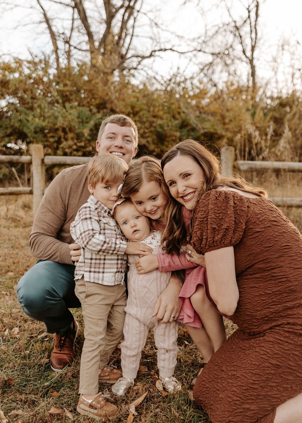 A happy family of five poses outdoors in fall, with parents and three young children smiling together near a wooden fence.