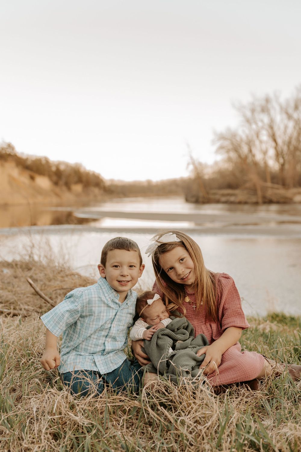 Two children sit by a river holding a newborn baby wrapped in a gray blanket on a fall day.