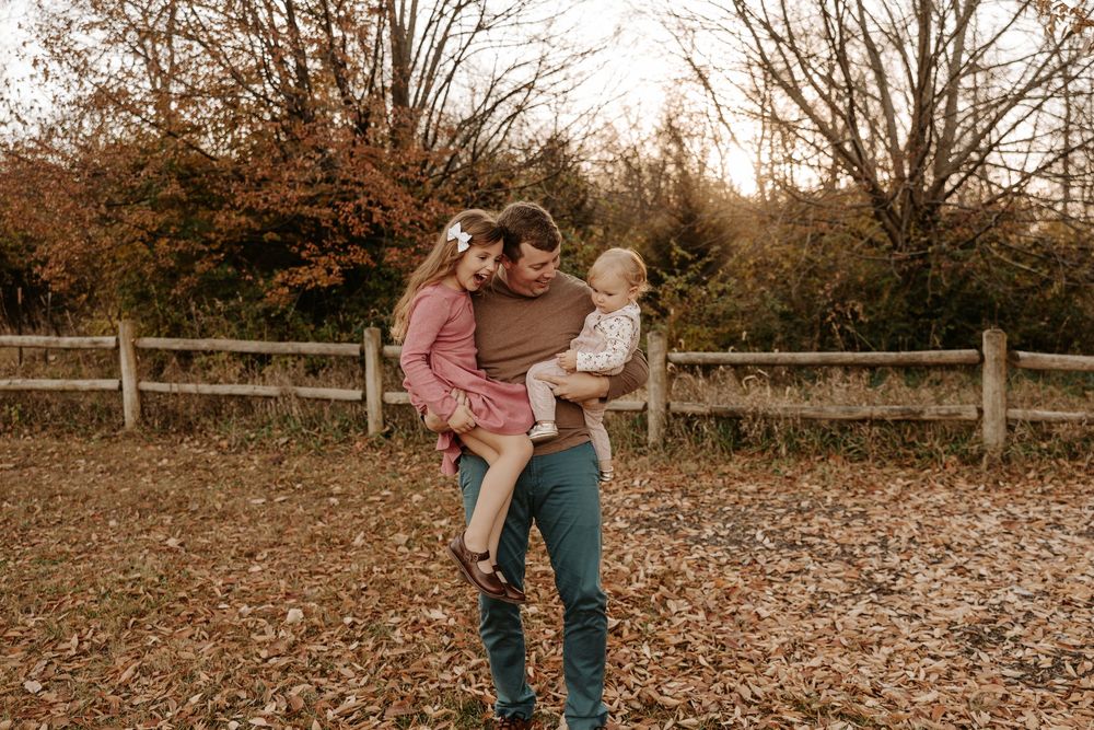 A father holds two young children in an autumn park with wooden fence and colorful fall foliage.
