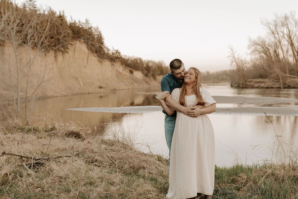 Couple embracing by a serene river with sandy cliffs and bare trees in a romantic outdoor engagement photo.