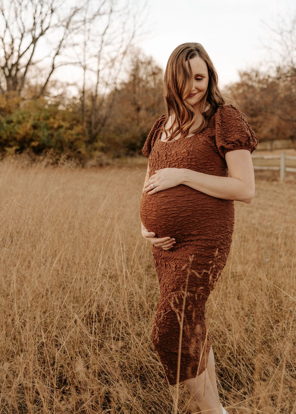 Pregnant woman in brown textured dress cradling her baby bump in a golden autumn field.
