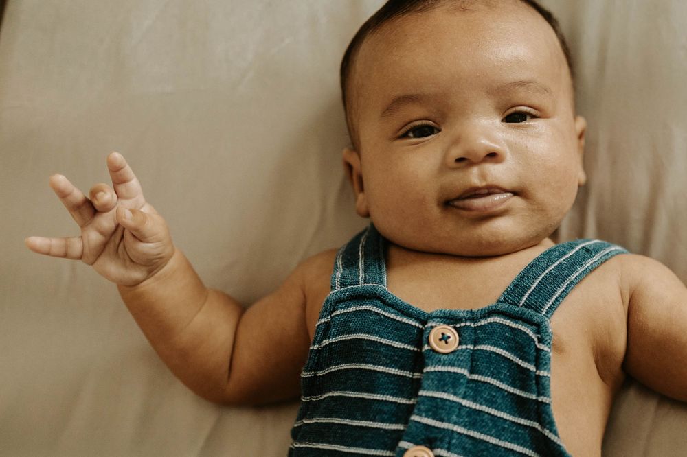 Baby in striped overalls waves at camera with a cheerful expression.