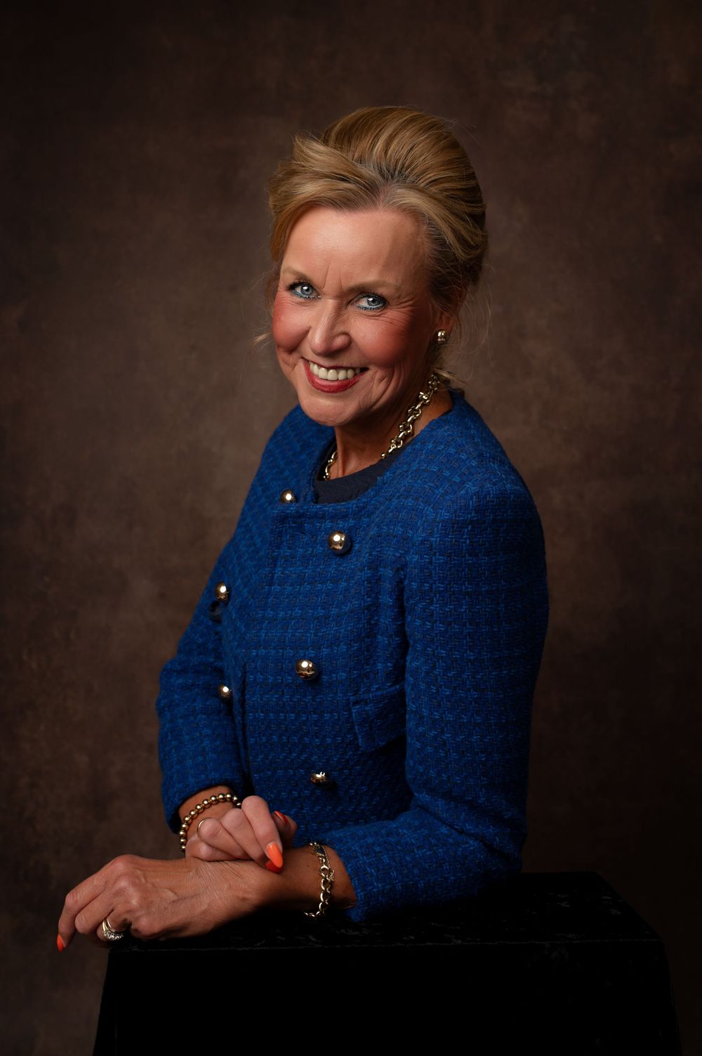 Professional studio portrait in royal blue sweater against dark brown backdrop with warm lighting and jewelry accents.