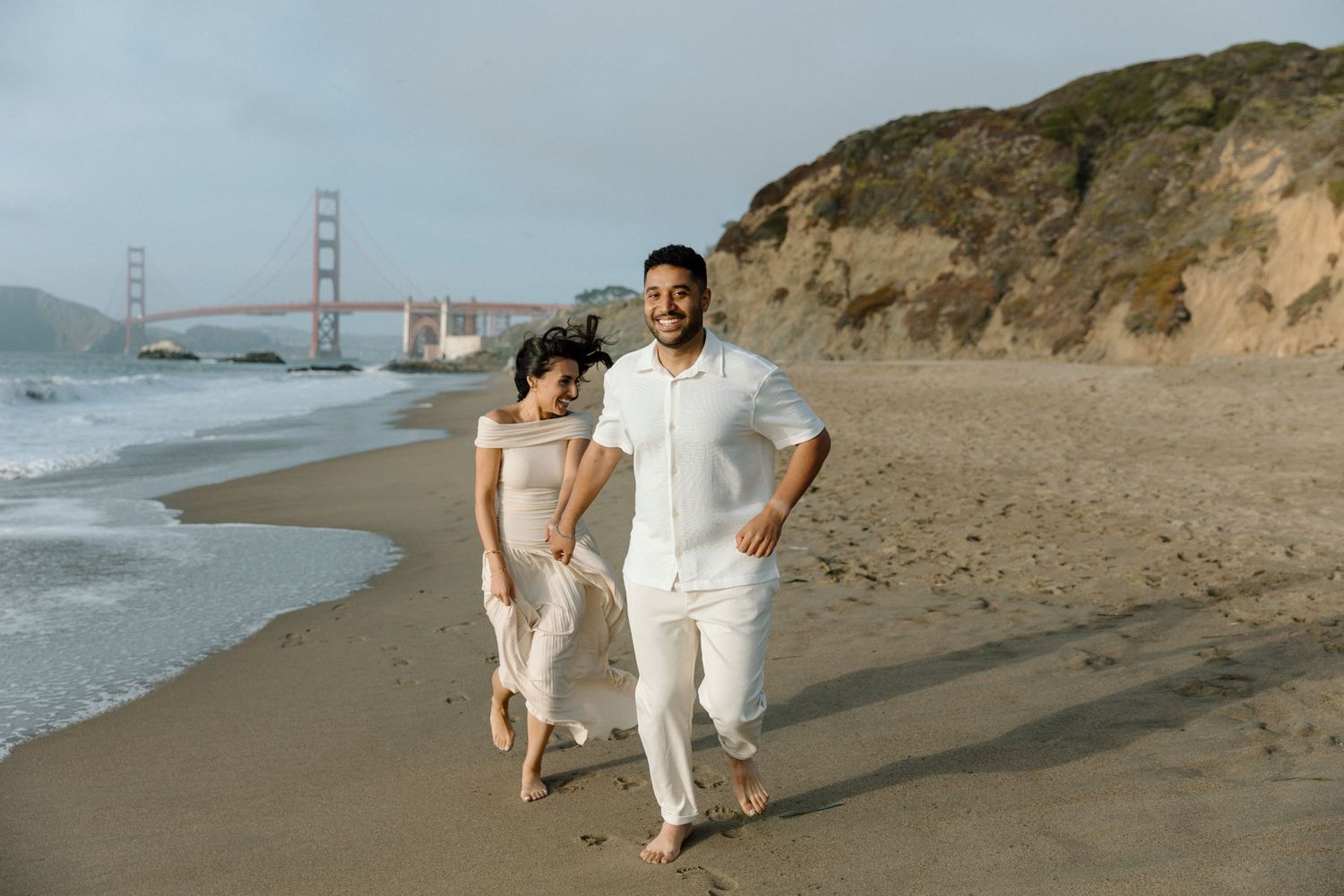 A couple strolls along Baker Beach with the Golden Gate Bridge in the background.