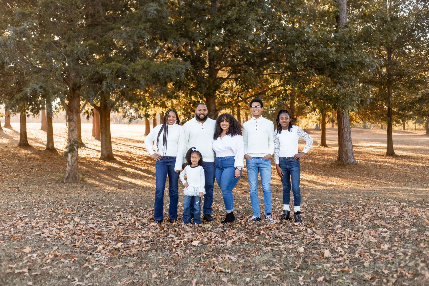 Family in coordinated white and denim outfits standing together among trees at sunset.