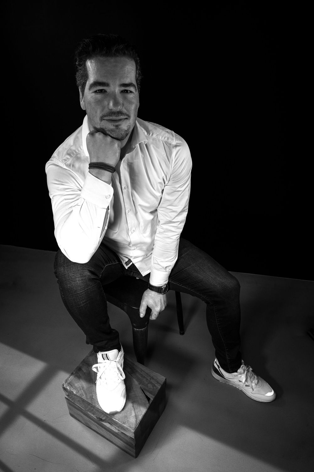 Black and white portrait of someone in white shirt and dark pants posing thoughtfully on chair against dark background.