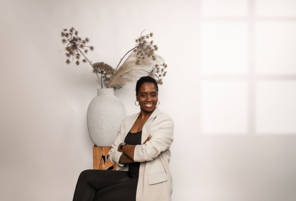 Subject wearing cream blazer poses next to modern vase with dried floral arrangement against white backdrop.