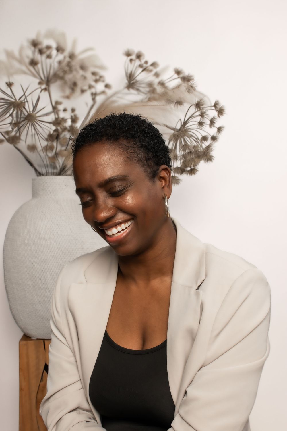 Person in beige blazer smiling warmly against neutral background with dried flowers in ceramic vase.