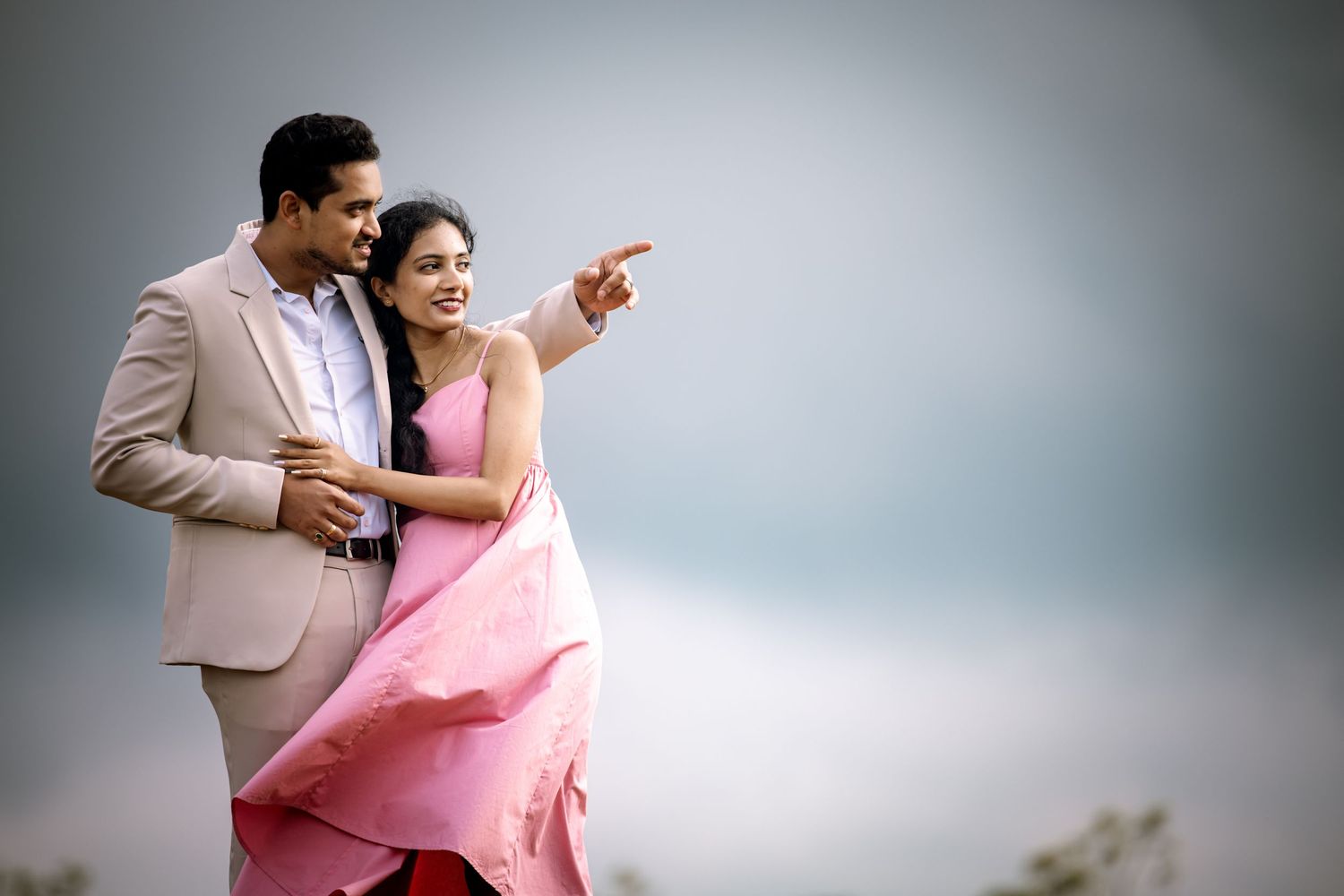 A romantic couple in pink and beige outfits embrace during an outdoor photoshoot.